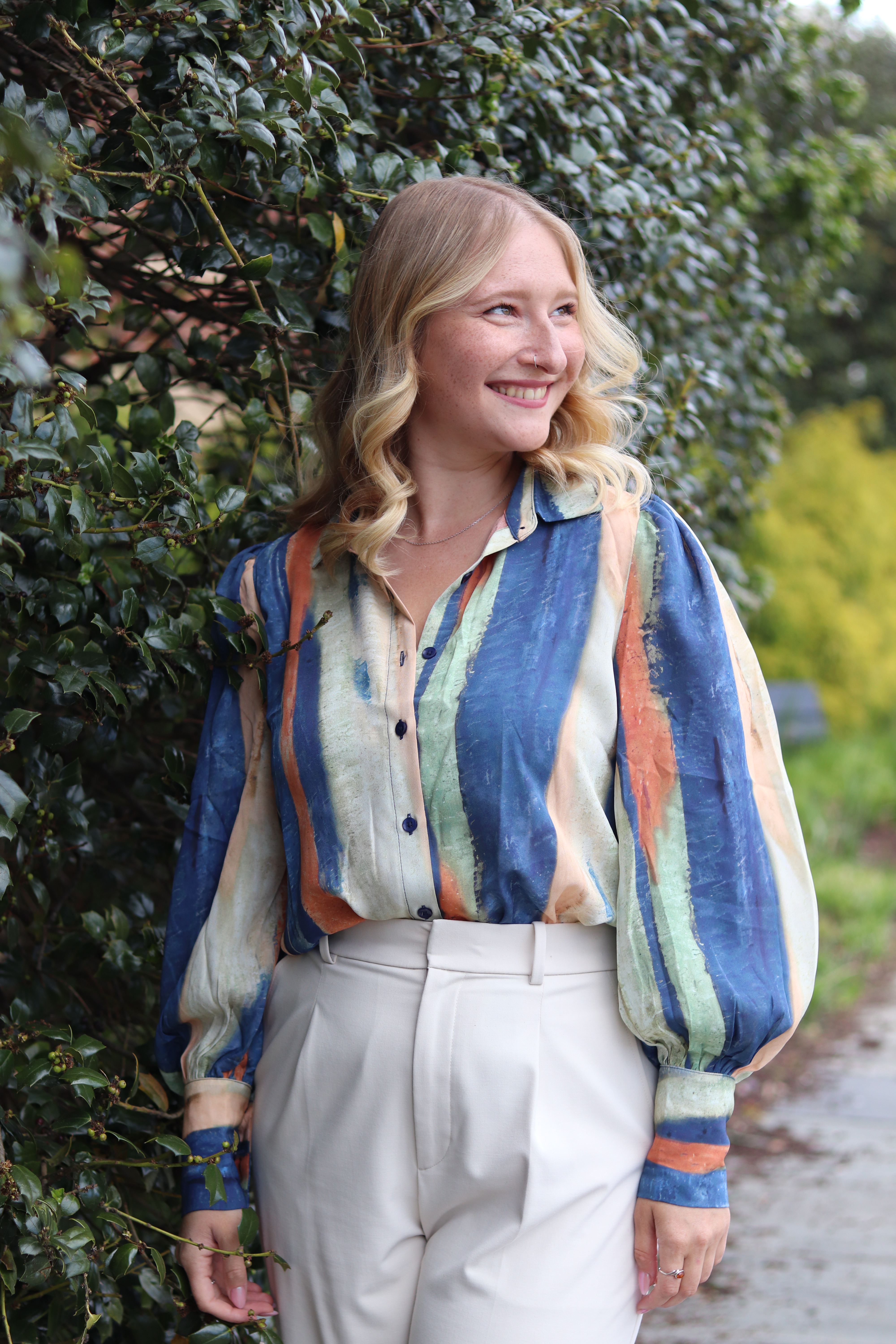 Alla walking past a green hedge in a painterly blouse and cream trousers, smiling softly.