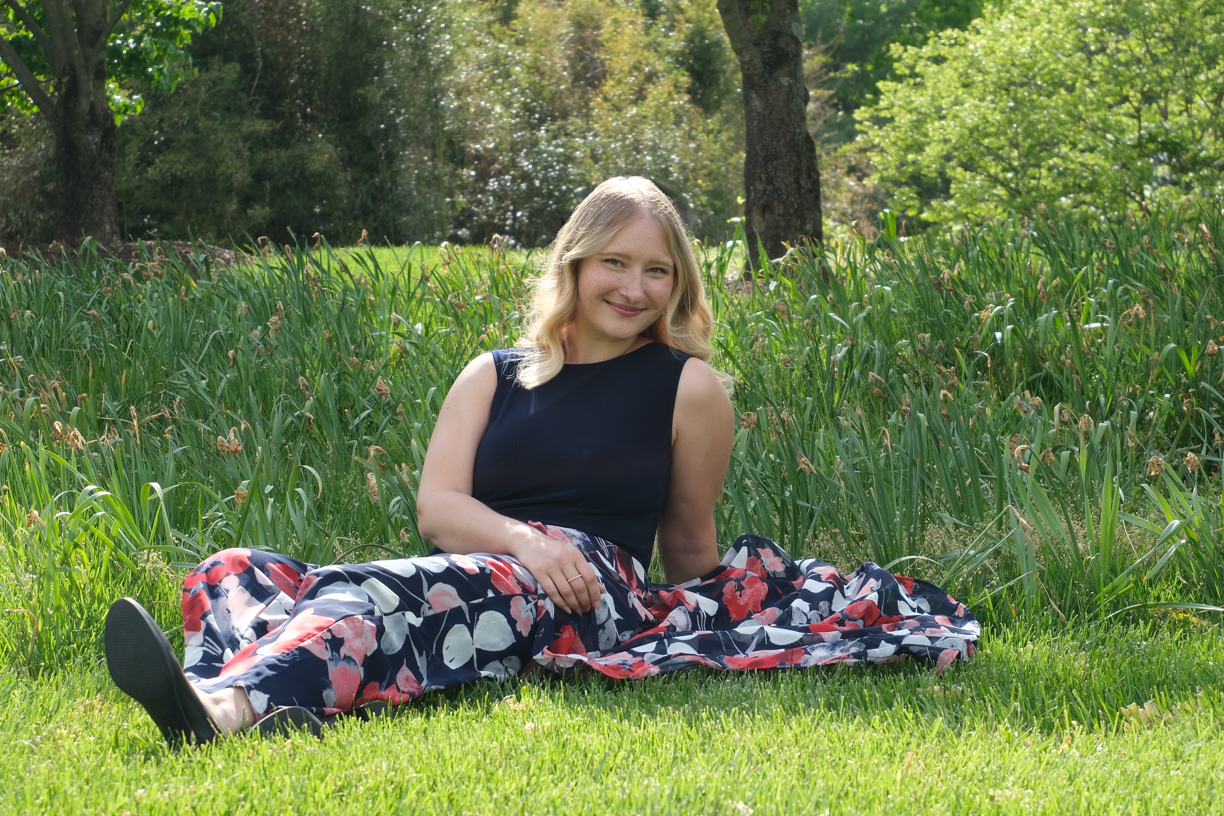Alla seated on a sunlit lawn in a navy top and floral skirt, smiling toward the camera.